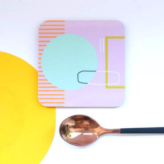 A square coaster with an abstract pink design, featuring geometric elements and negative space, alongside a copper-colored spoon on a yellow surface.