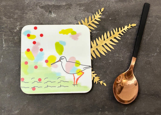 Coaster with a colorful bird design on a dark surface with a spoon and gold leaves.