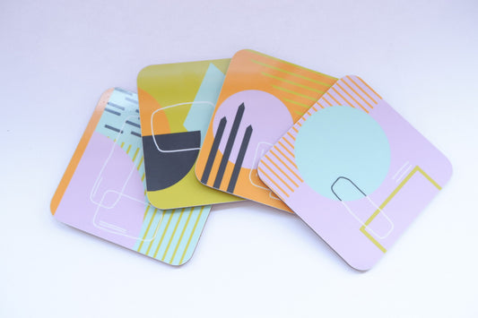 A set of four coasters with abstract geometric patterns in pink, blue, mustard, and orange colors.