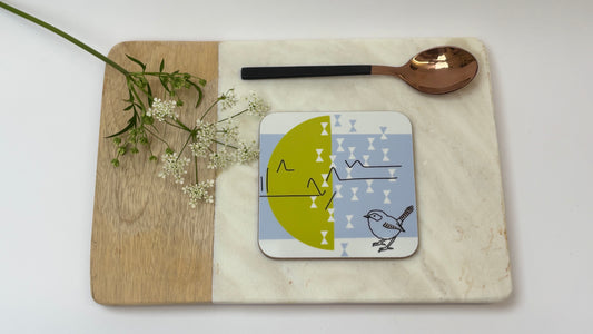 Coaster with bird design on a marble and wood surface with a spoon and flowers.