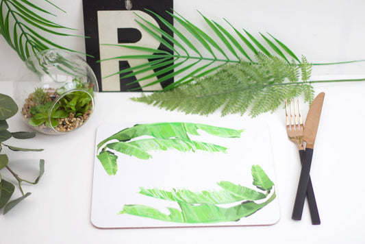 A green banana leaf placemat with a drawing of a palm tree in the background, placed on a white surface with a knife and a fork to the side.