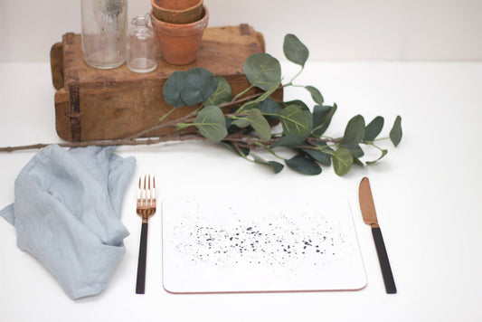 A black splatter placemat with a speckled pattern, displayed on a table with a blue napkin, black cutlery, a terracotta pot, and some green foliage for decoration.