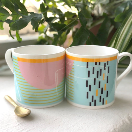 Two bone china mugs with abstract geometric painting in blue, pink, white, black, yellow, and orange colors, accompanied by a golden spoon, against a backdrop of green plants.