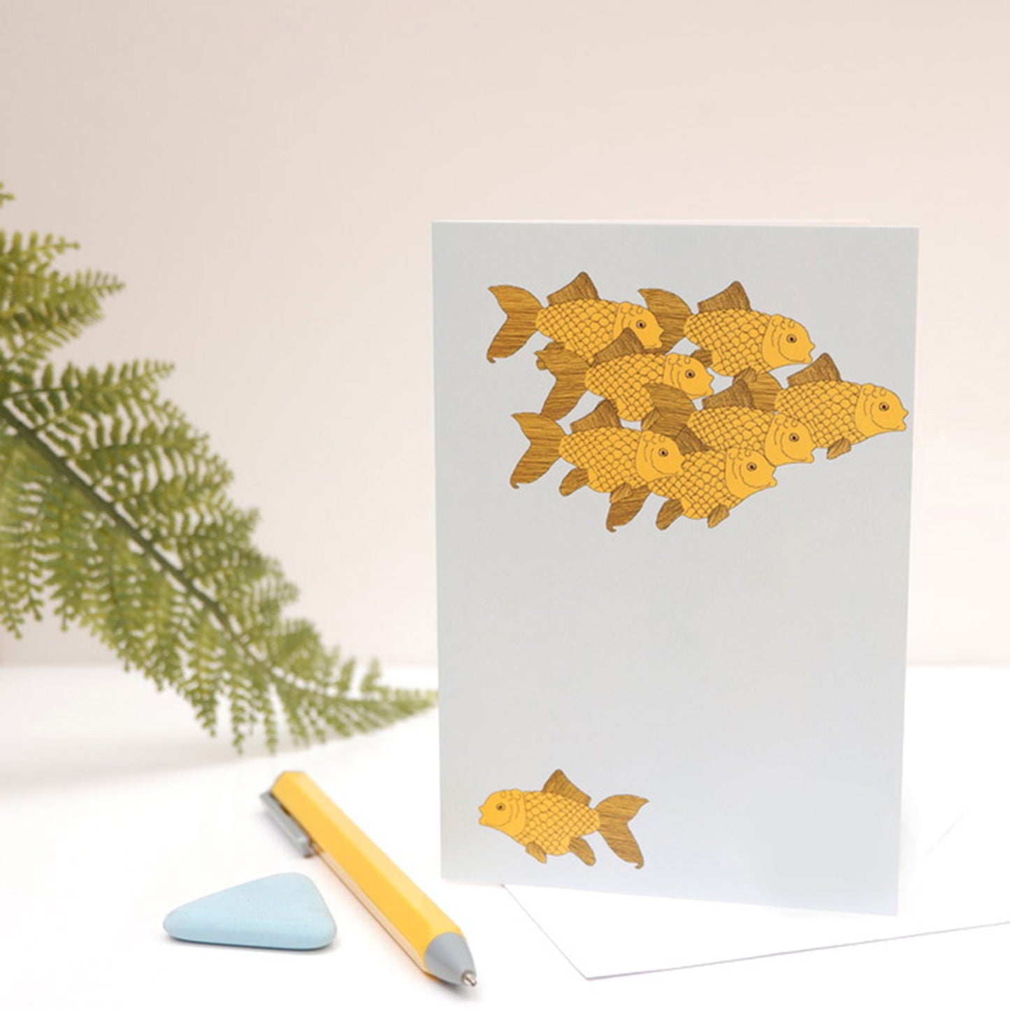 A greetings card with an illustration of goldfish swimming against a blue background, placed on a white surface with a pencil and a green leaf to the side.