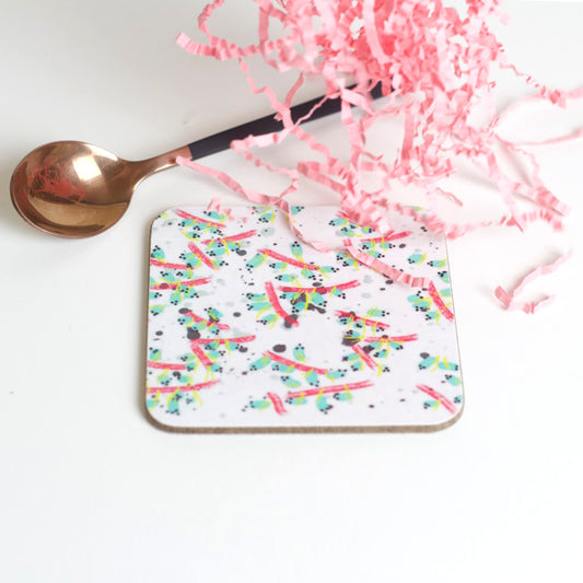A square coaster with a white background featuring a multicolored, abstract pattern of lines and shapes, placed on a white surface next to a bronze spoon and pink tissue paper.