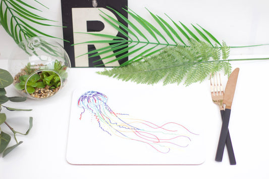 A placemat with a jellyfish design, part of the underwater collection, displayed on a table with a plant, a coaster, and a brush.