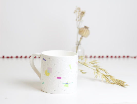 A white bone china mug with multicolored patterns and a fine rim, placed on a surface with dried flowers and a decorative red ribbon in the background.