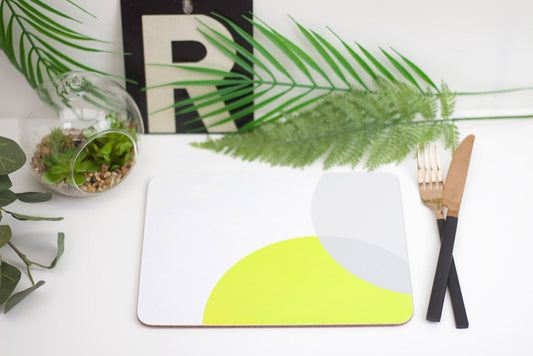 A lime green and grey placemat with a geometric design, overlaid on a white surface, with a wooden cutlery set and a potted plant in the background.