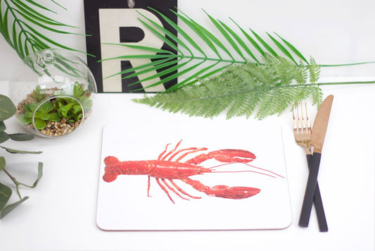 A placemat with a red lobster design, a set of utensils on a white surface with a glass vase with green leaves in the background.