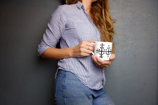 A person holding a bone china mug with a geometric pattern, specifically a cross, on a blue and white striped background.
