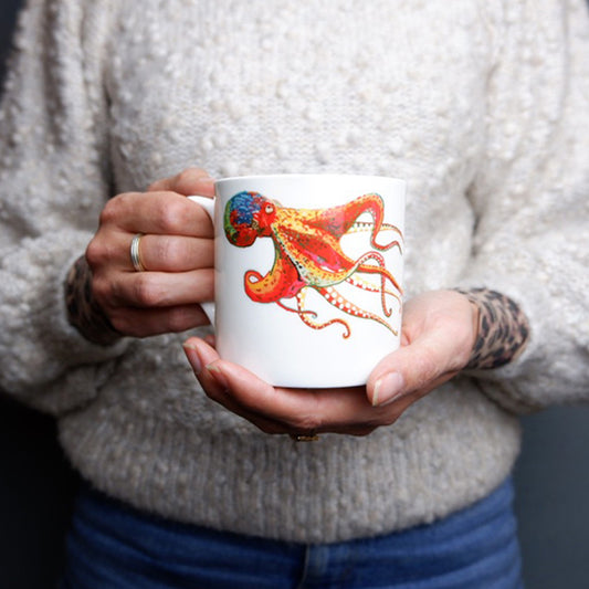 A person holding a white bone china mug with a colorful hand-drawn Octopus design on it.