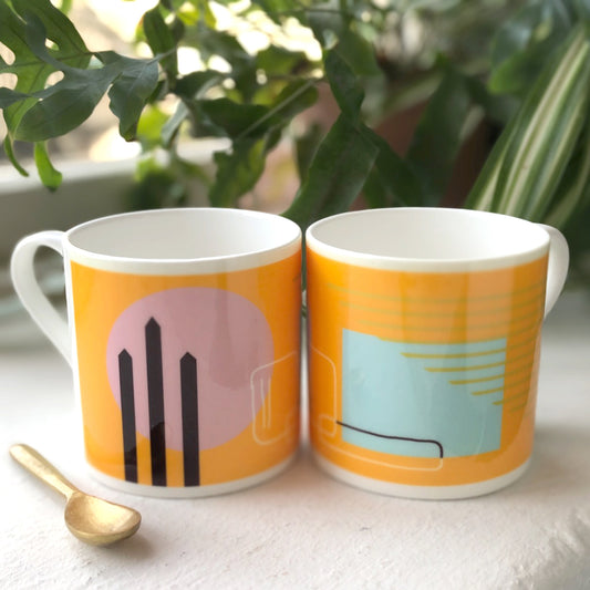 Two bone china mugs with abstract geometric paintings in orange, pink, and blue colors, placed on a surface with a golden spoon beside them.