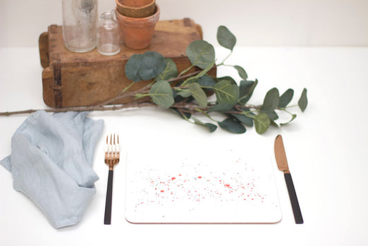 A splatter placemat in orange with multicolored specks, placed on a white surface with a blue napkin, a fork, and a potted plant in the background.