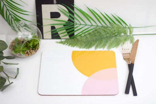 A placemat with a geometric design featuring orange and pink shades, placed on a table with a plant, a glass, and a wooden fork.