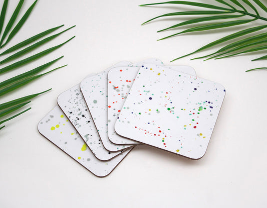 A set of four square coasters with a white base and multicolored splatter design, displayed on a white surface.