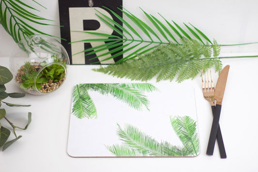 A placemat with a palm leaf design, laid out on a table with a green plant and a wooden cutlery set on top of it.