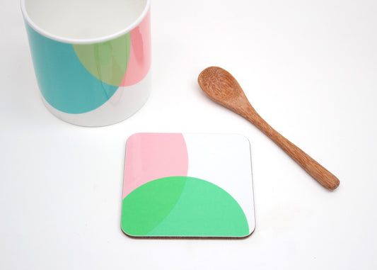 A coaster with a pink and green geometric pattern, a bone china mug with a geometric pattern, and a wooden spoon on a white background.