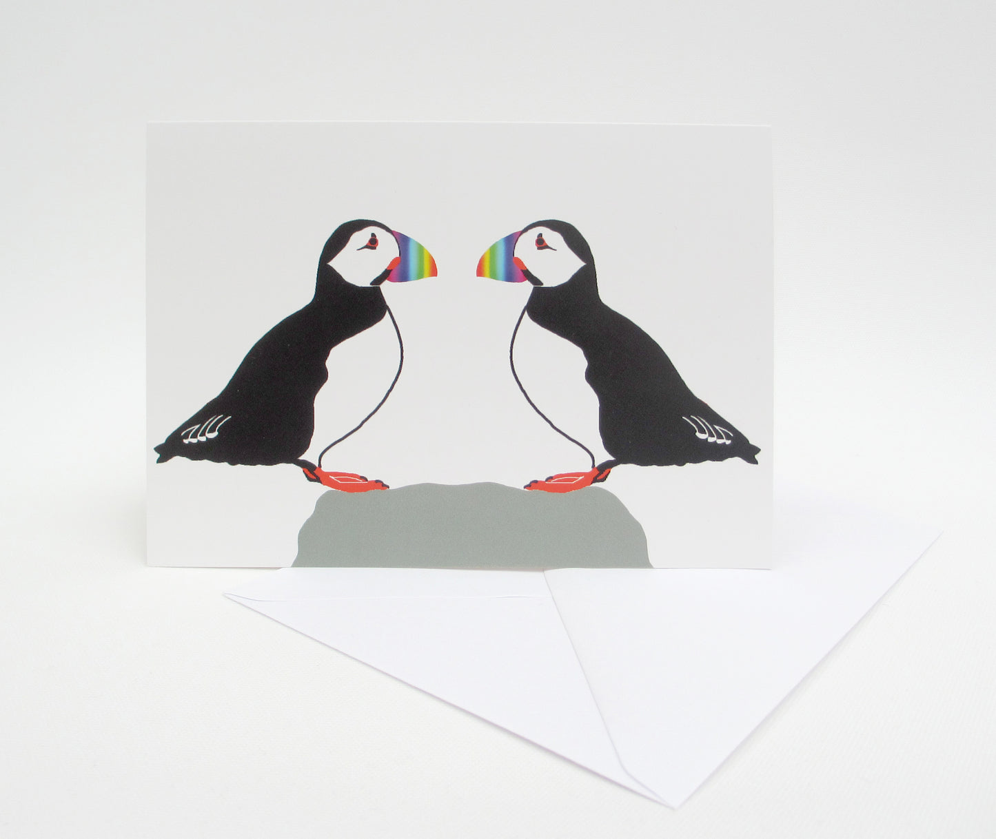 A greeting card with a illustration of two puffins facing each other. The card is set against a white background and there is a white envelope included.