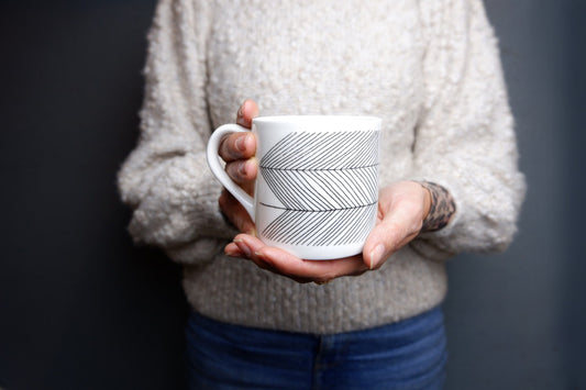 A person holding a white bone china mug with a geometric pattern.