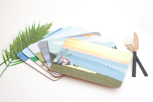 A collection of colorful placemats with various designs, including a beach scene and a forest, displayed on a table with a fork and a green plant in the background.