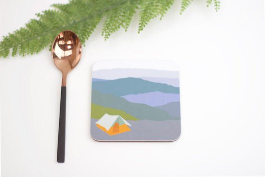 A square coaster with a camping tent design in the Picos mountains, accompanied by a copper-colored spoon and green foliage in the background.
