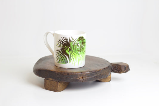 A bone china mug with a design of tropical leaves, specifically cabbage, placed on a wooden stand against a white background.