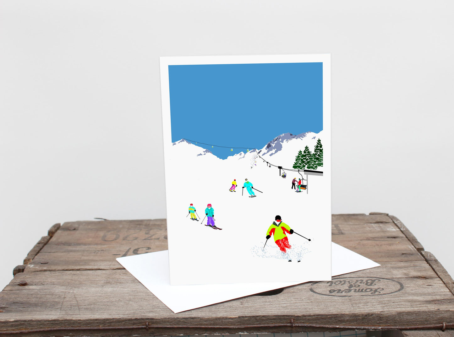 A greetings card with a winter skiing scene, featuring mountains, skiers, and trees on a blue background.