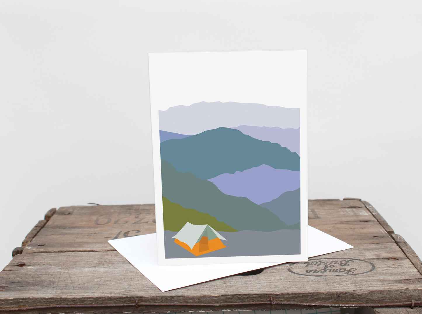 A greetings card with a mountain range and a tent design, placed on a wooden surface.