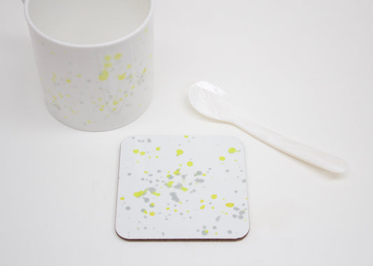 A white coaster with yellow splatter design, placed on a white surface with a cup and a white spoon next to it.