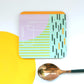 A set of square coasters with abstract blue and orange geometric patterns, one coaster is placed under a spoon to show the design.