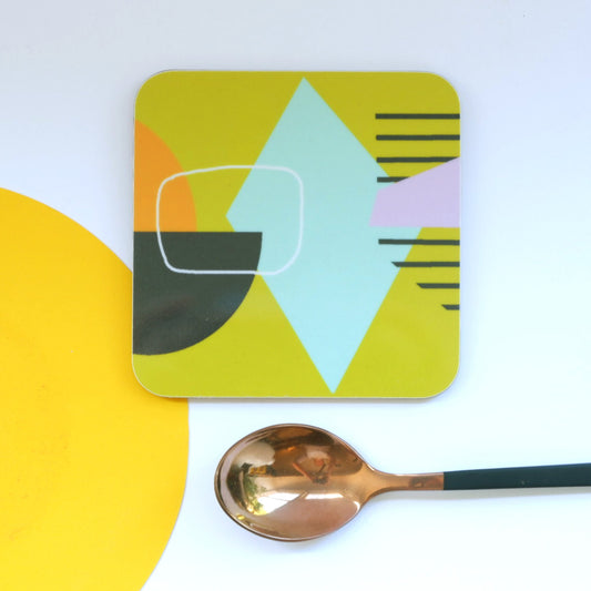 A square coaster with an abstract mustard-colored design, placed on a white surface next to a spoon for scale.