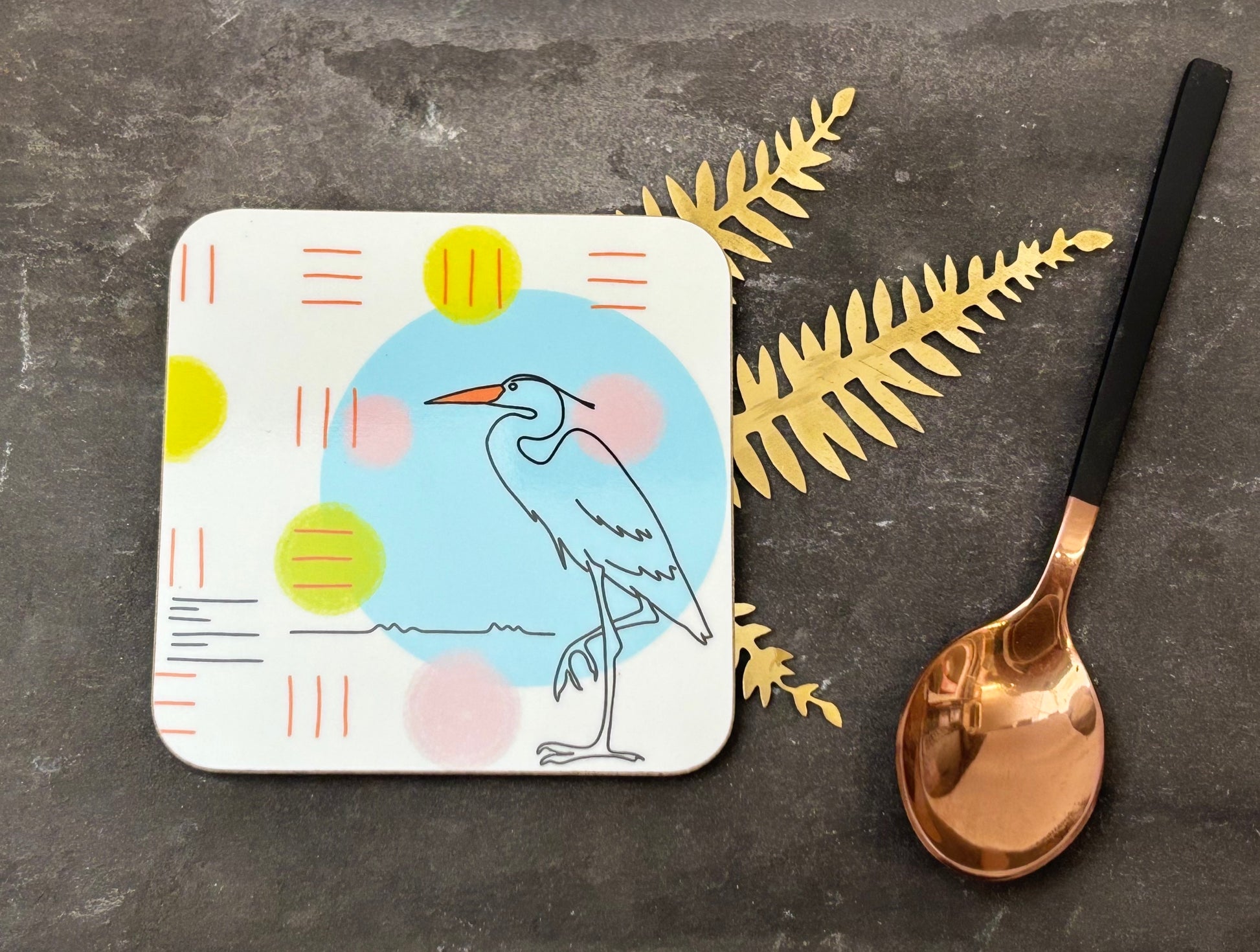Coaster with bird design on a dark surface with a spoon and decorative leaves