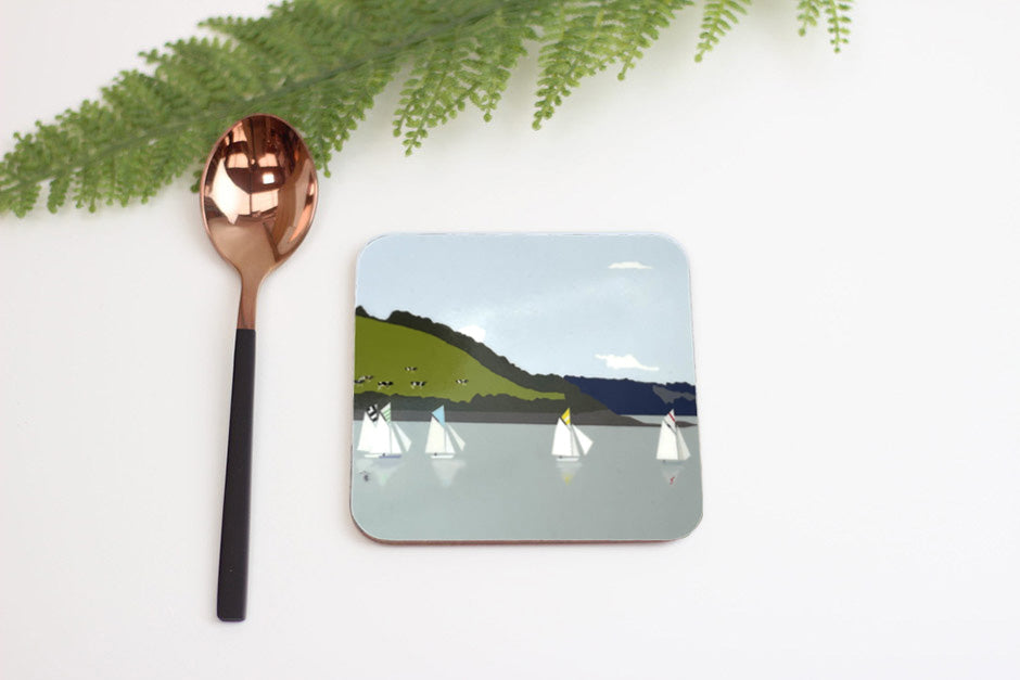 Coaster with sailboat design on a white surface with a copper spoon and greenery.