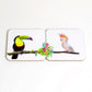A set of two square coasters with hand-drawn illustrations of a toucan and a cockatoo on a jungle-themed background, placed on a white wall.