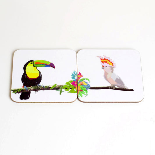 A set of two square coasters with hand-drawn illustrations of a toucan and a cockatoo on a jungle-themed background, placed on a white wall.