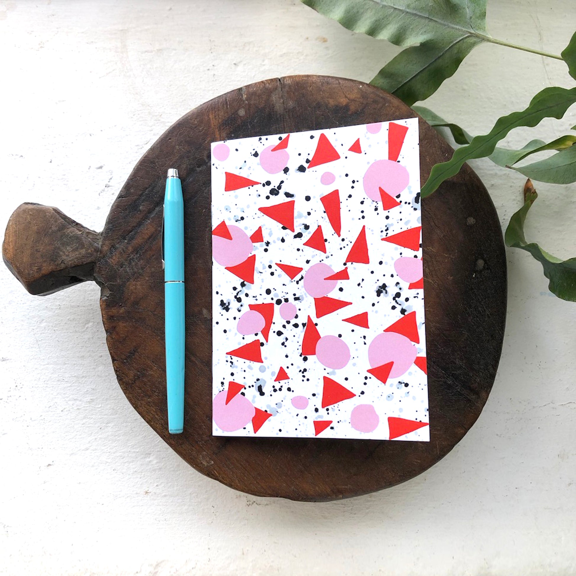 A6 notebook with a geometric pattern in red and white colors, placed on a wooden board next to a blue pen.