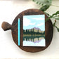 A6 notebook with a landscape design on the cover, featuring a blue sky, green trees, and a mountain in the background, placed on a wooden round panel next to a blue pen.
