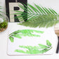 A green banana leaf placemat with a drawing of a palm tree in the background, placed on a white surface with a knife and a fork to the side.