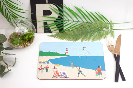 A colorful beach-themed placemat with a design that includes beach chairs, an umbrella, and a fishing line, positioned on a table with a fork and a knife to the side.