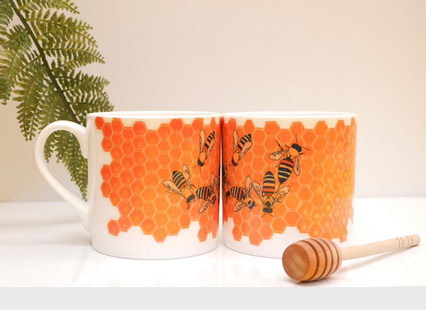 A bone china mug with a orange geometric honeycomb pattern and bees on it, displayed on a white surface with a plant in the background and a wooden spoon to the side.