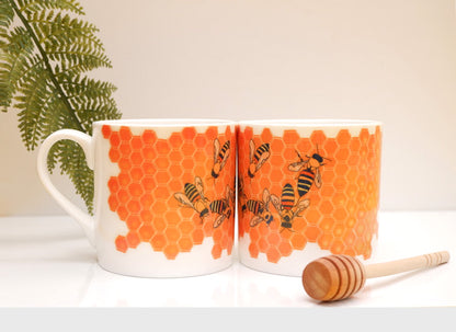 A bone china mug with a orange geometric honeycomb pattern and bees on it, displayed on a white surface with a plant in the background and a wooden spoon to the side.