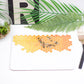 A placemat with a geometric honeycomb pattern and bee illustrations in orange shades, displayed on a table with cutlery and decorative plants around it.