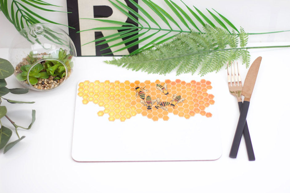 A placemat with a geometric honeycomb pattern and bee illustrations in orange shades, displayed on a table with cutlery and decorative plants around it.