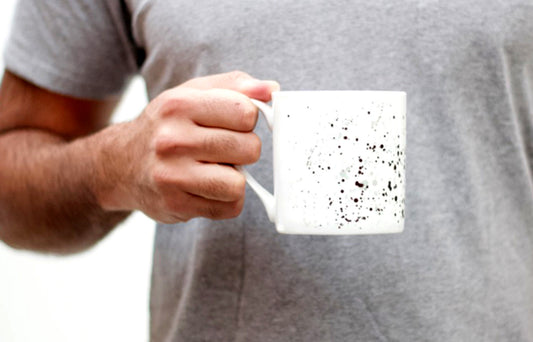 A person is holding a white bone china mug with black and grey speckled pattern.