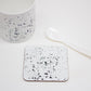 A white coaster with black paint splatter design, displayed alongside a white mug and a spoon on a white surface.