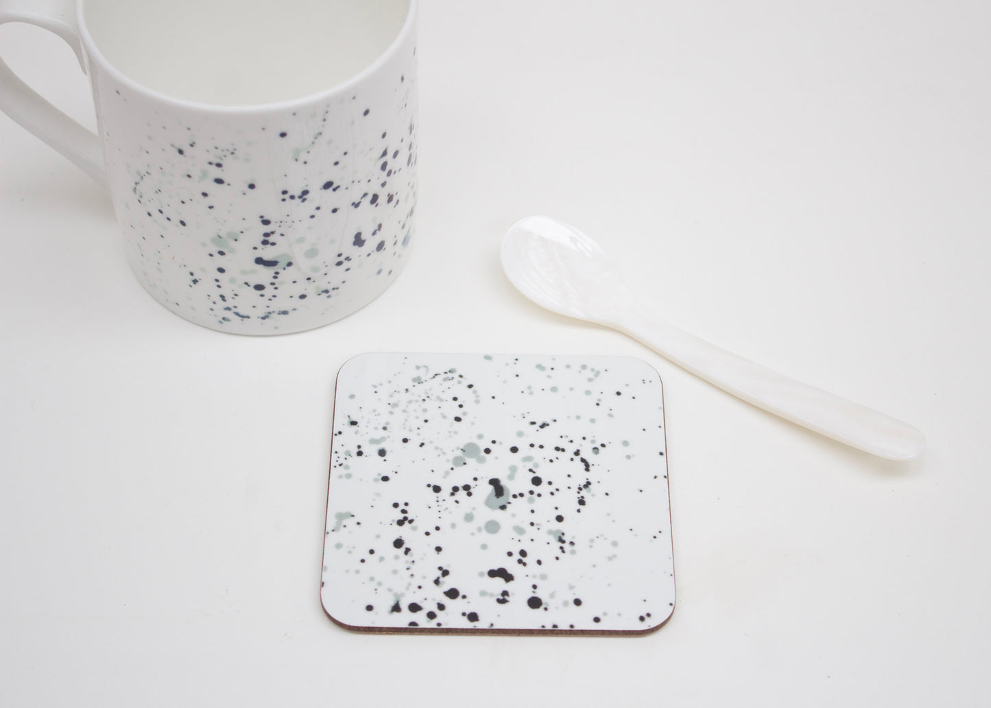 A white coaster with black paint splatter design, displayed alongside a white mug and a spoon on a white surface.
