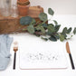 A black splatter placemat with a speckled pattern, displayed on a table with a blue napkin, black cutlery, a terracotta pot, and some green foliage for decoration.
