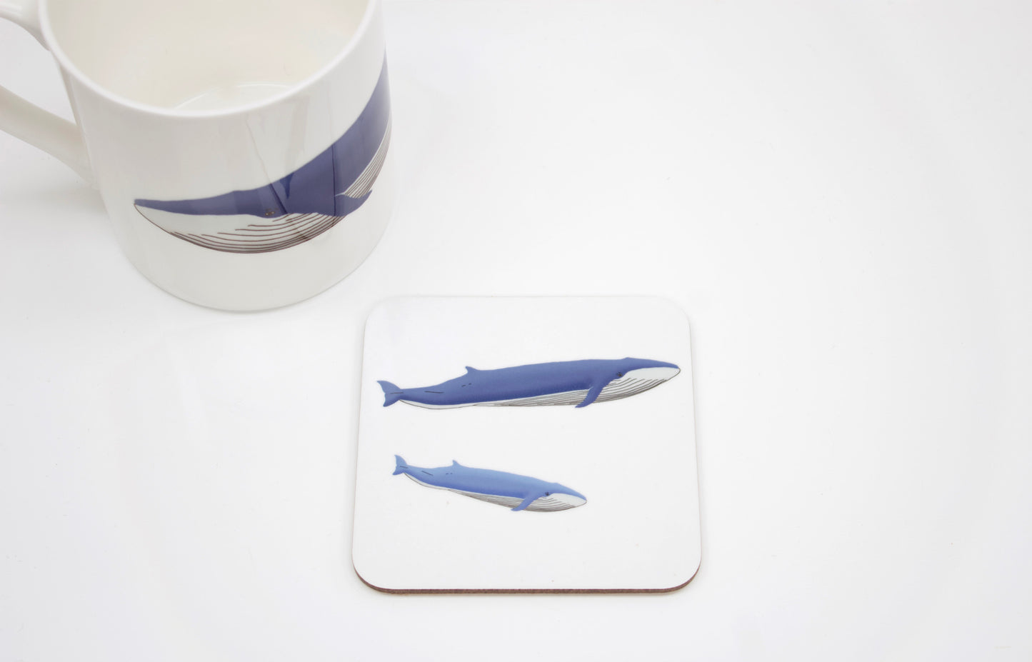 A square coaster with a cork base featuring a hand-drawn design of a blue whale and her calf, accompanied by a white mug with a similar design.