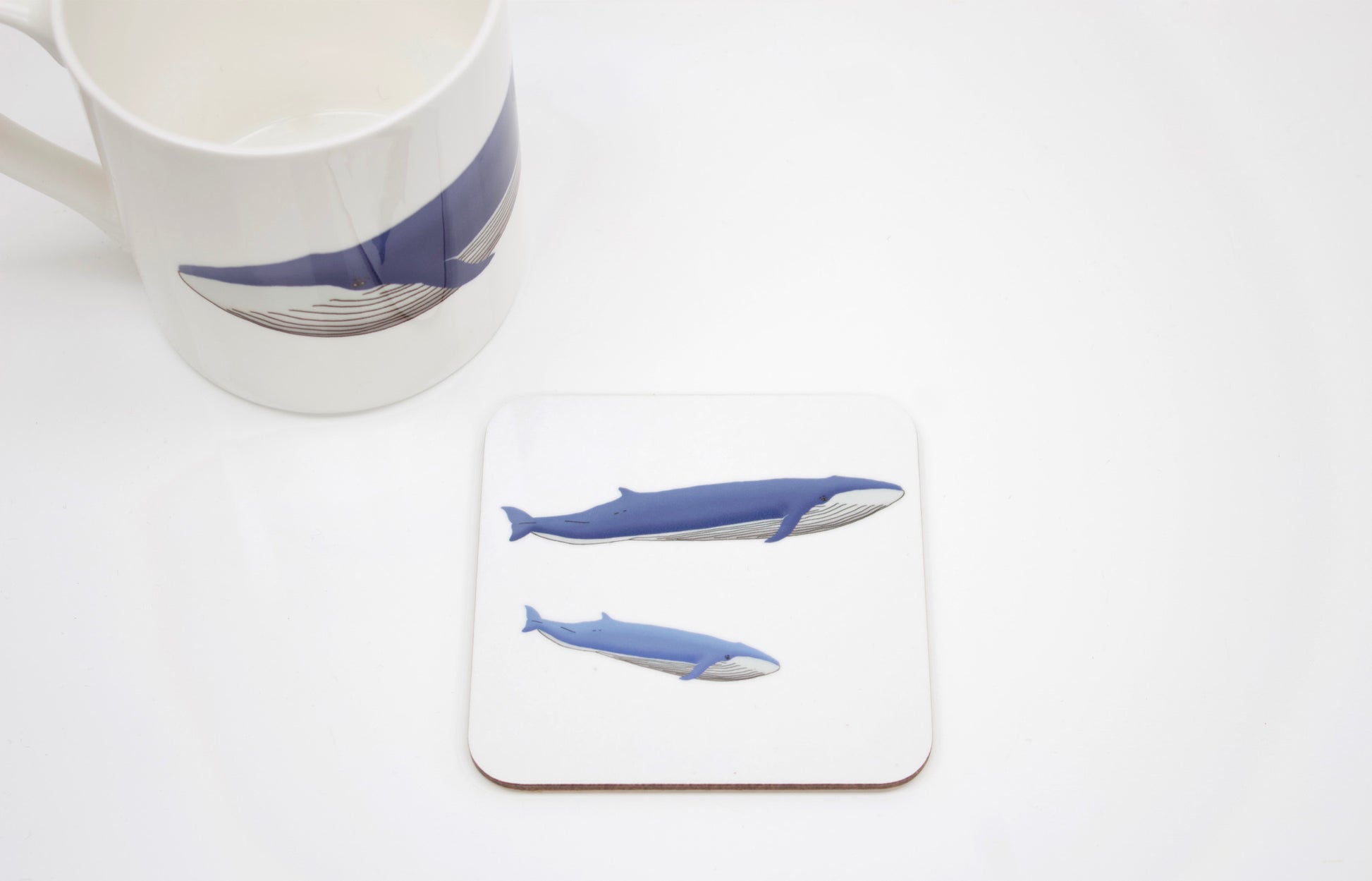 A square coaster with a cork base featuring a hand-drawn design of a blue whale and her calf, accompanied by a white mug with a similar design.