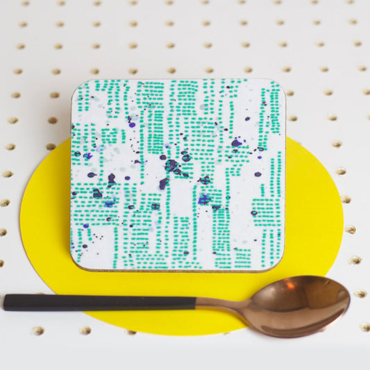 A square coaster with a blurred pattern of ocean green dots, blue and grey splashes, placed on a yellow plate with a spoon next to it, against a white background with pins.